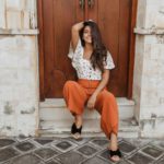 Charming curly brunette woman in culottes and white blouse sits on threshold of house with wooden d