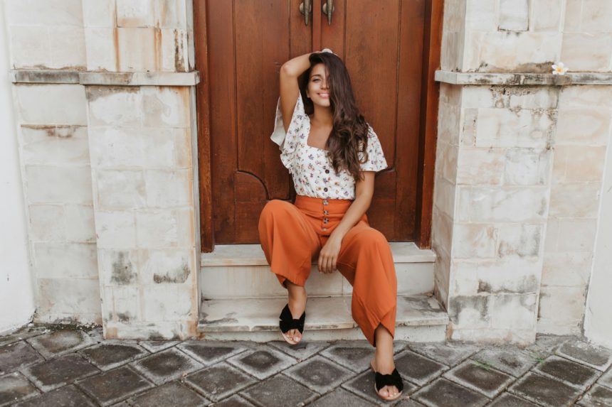 Charming curly brunette woman in culottes and white blouse sits on threshold of house with wooden d