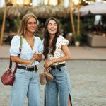 Young beautiful girlfriends in denim pants and trendy floral colorful blouses smile and look away.