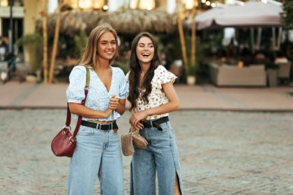 Young beautiful girlfriends in denim pants and trendy floral colorful blouses smile and look away.